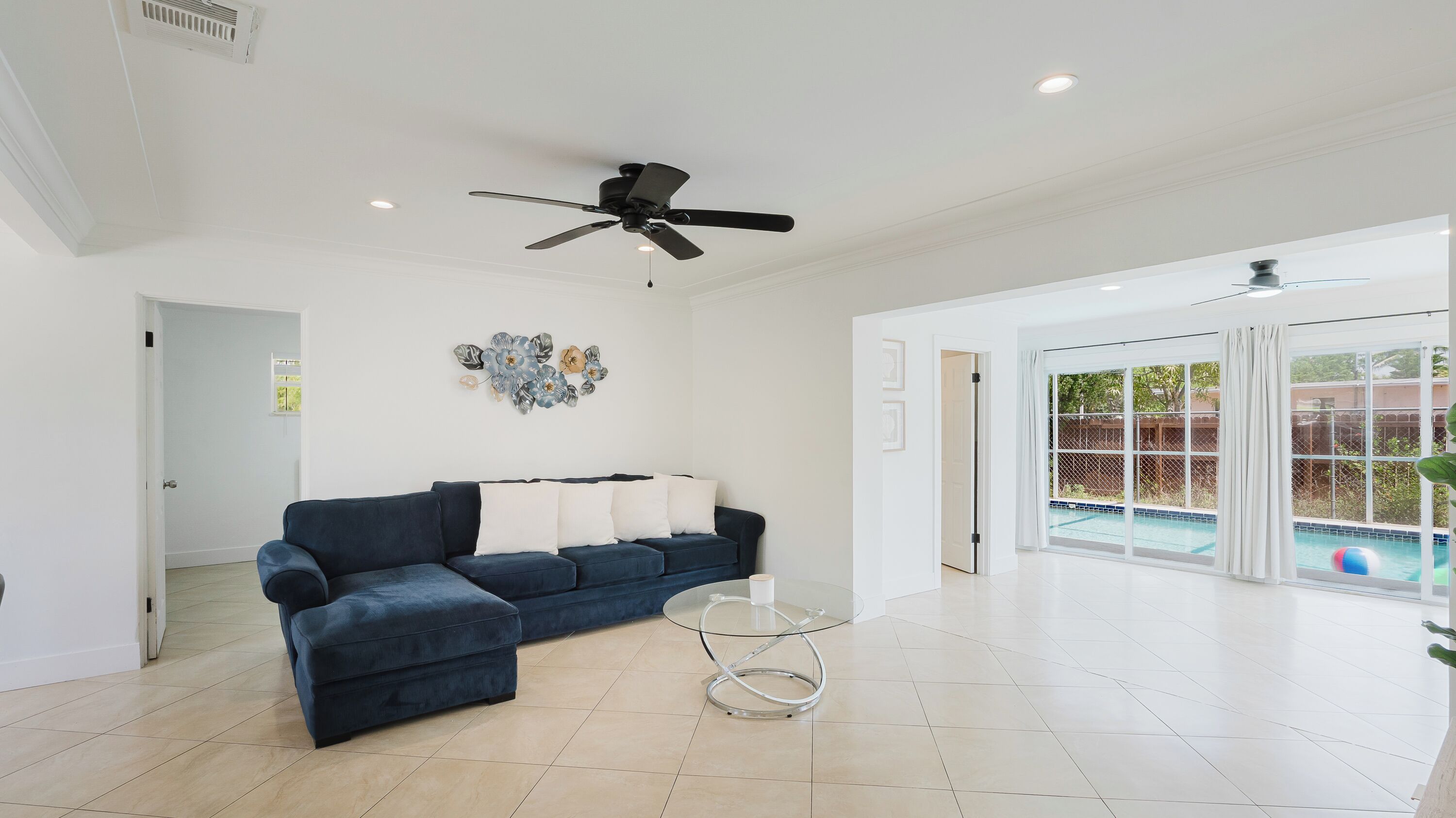 A spacious living room with a dark blue sofa, a glass coffee table, a ceiling fan, and a view of an outdoor swimming pool through large sliding glass doors.