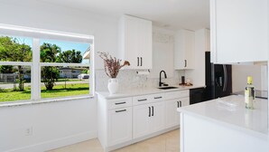 A modern kitchen with white cabinetry, a black refrigerator, and a window with a view of a green lawn and trees. - FLORIDA PARADISE / 5 MIN TO DOWNTOWN / 3BDR/6BEDS (Fort Myers)