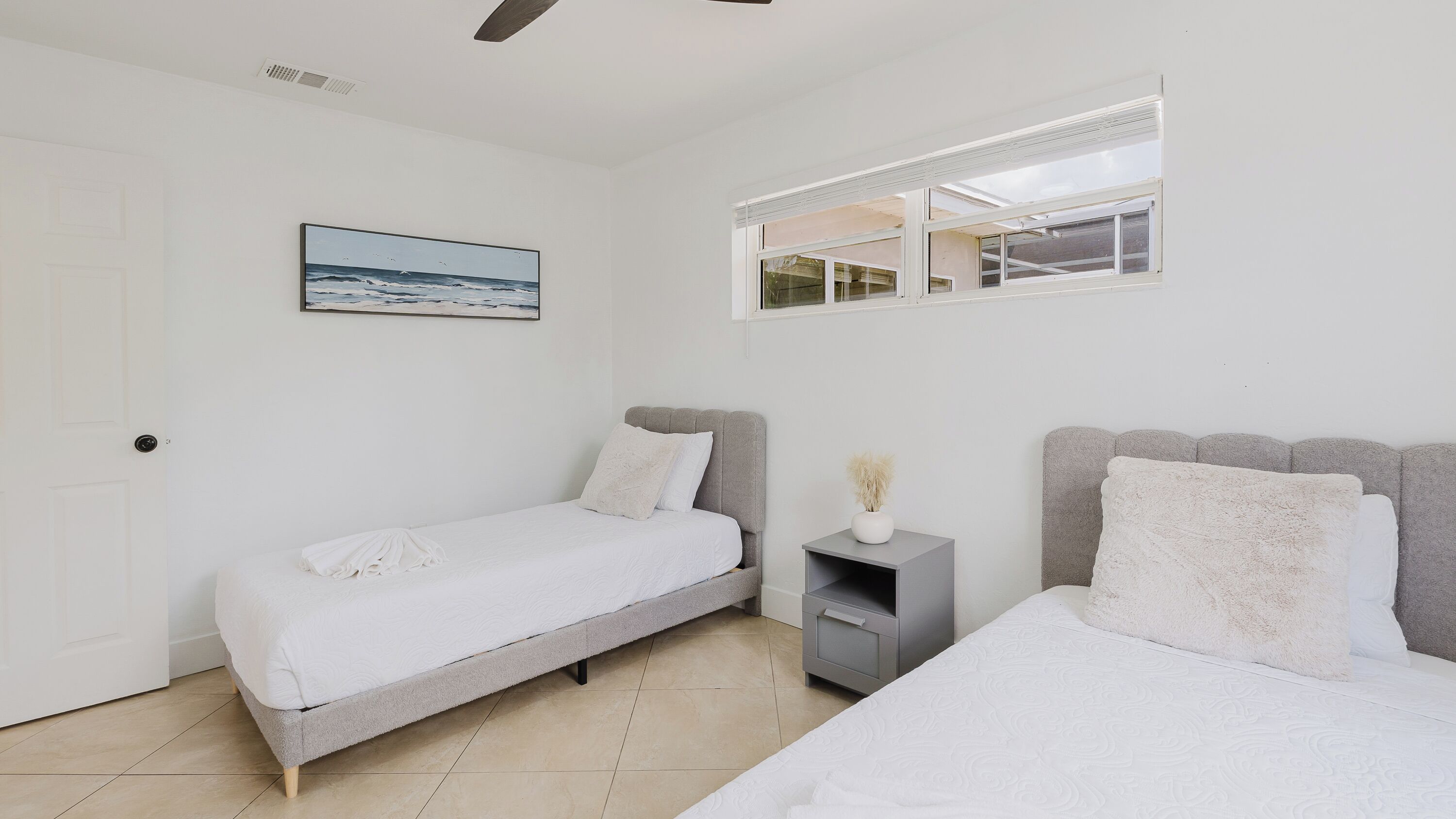A bedroom with two beds, a window, a painting, and a nightstand.