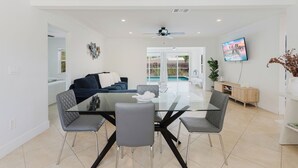 A modern living room with a glass dining table, grey upholstered chairs, a blue sofa, and a television mounted on the wall. - FLORIDA PARADISE / 5 MIN TO DOWNTOWN / 3BDR/6BEDS (Fort Myers)