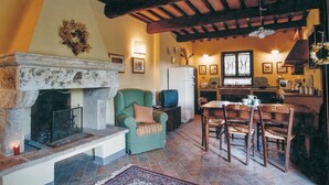A cozy living room with a fireplace, a dining area with wooden chairs, and a kitchen with a refrigerator and window.