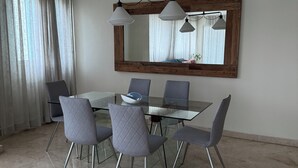A dining area with a glass table surrounded by six chairs, a large mirror above the table, and a chandelier with three lamps.
