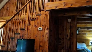 A wooden cabin interior with a wooden staircase, a television on a wooden stand, and a bed in the adjacent room.