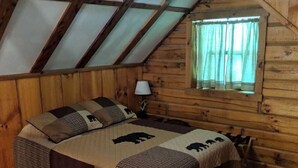 A cozy wooden cabin room with a sloped ceiling, a bed with a quilted blanket, a nightstand, and a window with curtains.