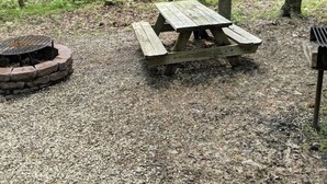 A wooden picnic table surrounded by a forest with a fire pit nearby.