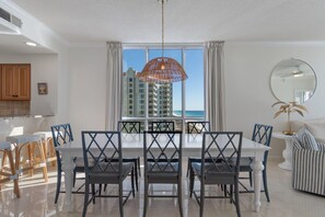 A dining area with a white table and chairs, a large window with a view of a high-rise building and the ocean, and a ceiling fan. - Dog Friendly, Gulf Front Condo-Heated Pool/Hot tub (Navarre Beach)