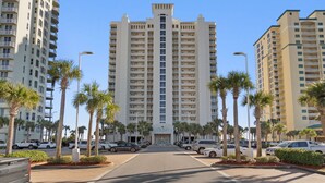 A tall hotel with multiple floors, a central entrance, and a parking area with cars and palm trees. - Luxurious Gulf-front Condo, Heated Pool, Spa/Gym (Navarre Beach)