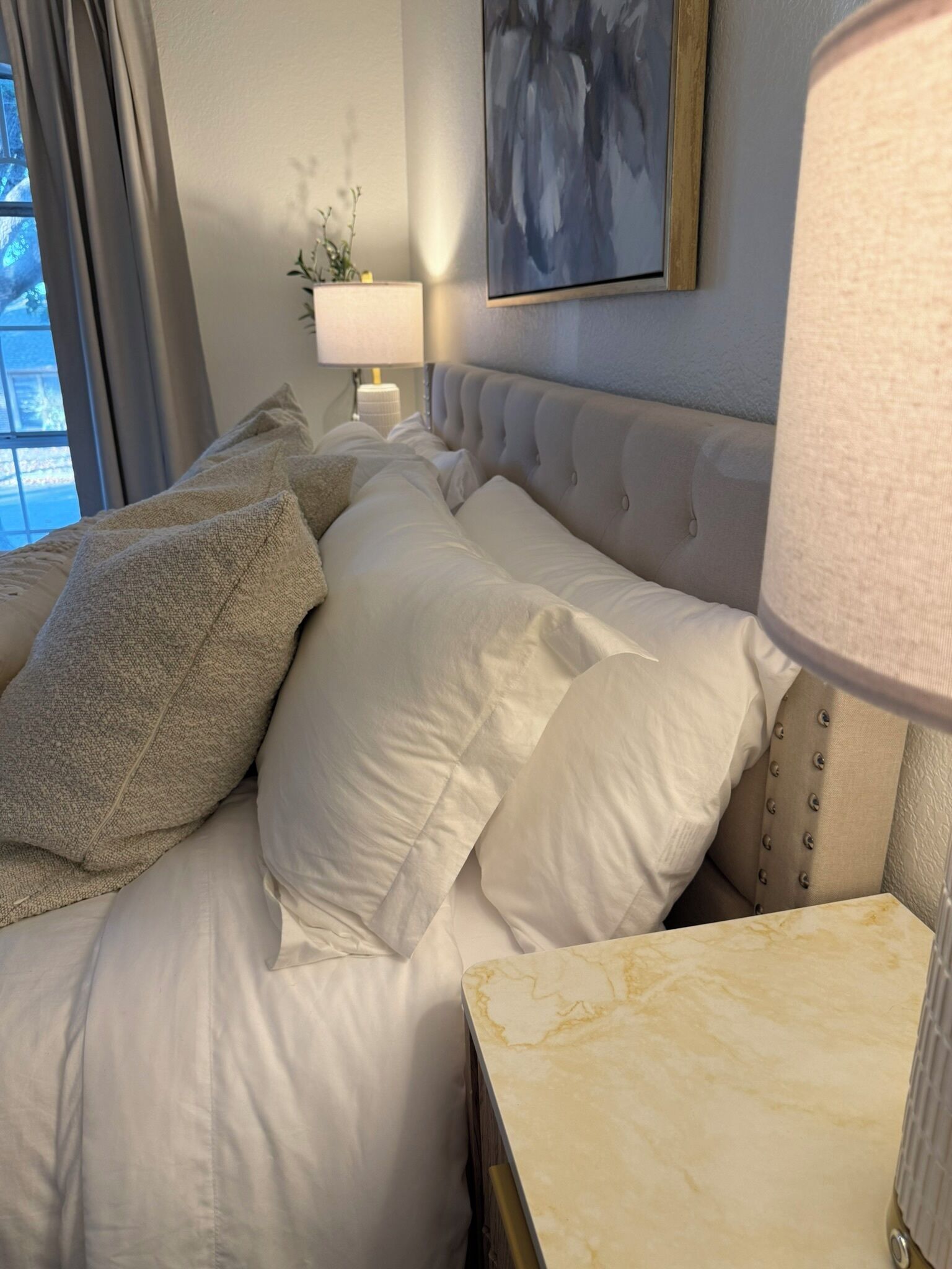 Una cama bien hecha con sábanas blancas y varios almohadones, una mesita de noche de mármol, una lámpara y un cuadro en la pared.