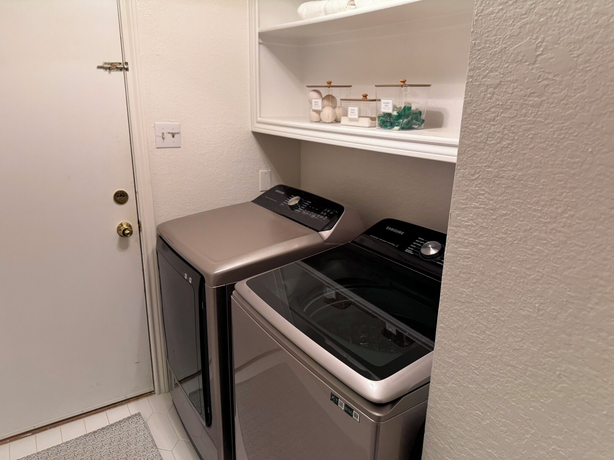 Un espace réduit dédié à la buanderie, comprenant un lave-linge et un sèche-linge, une petite étagère pour les articles de toilette et une porte avec une poignée.