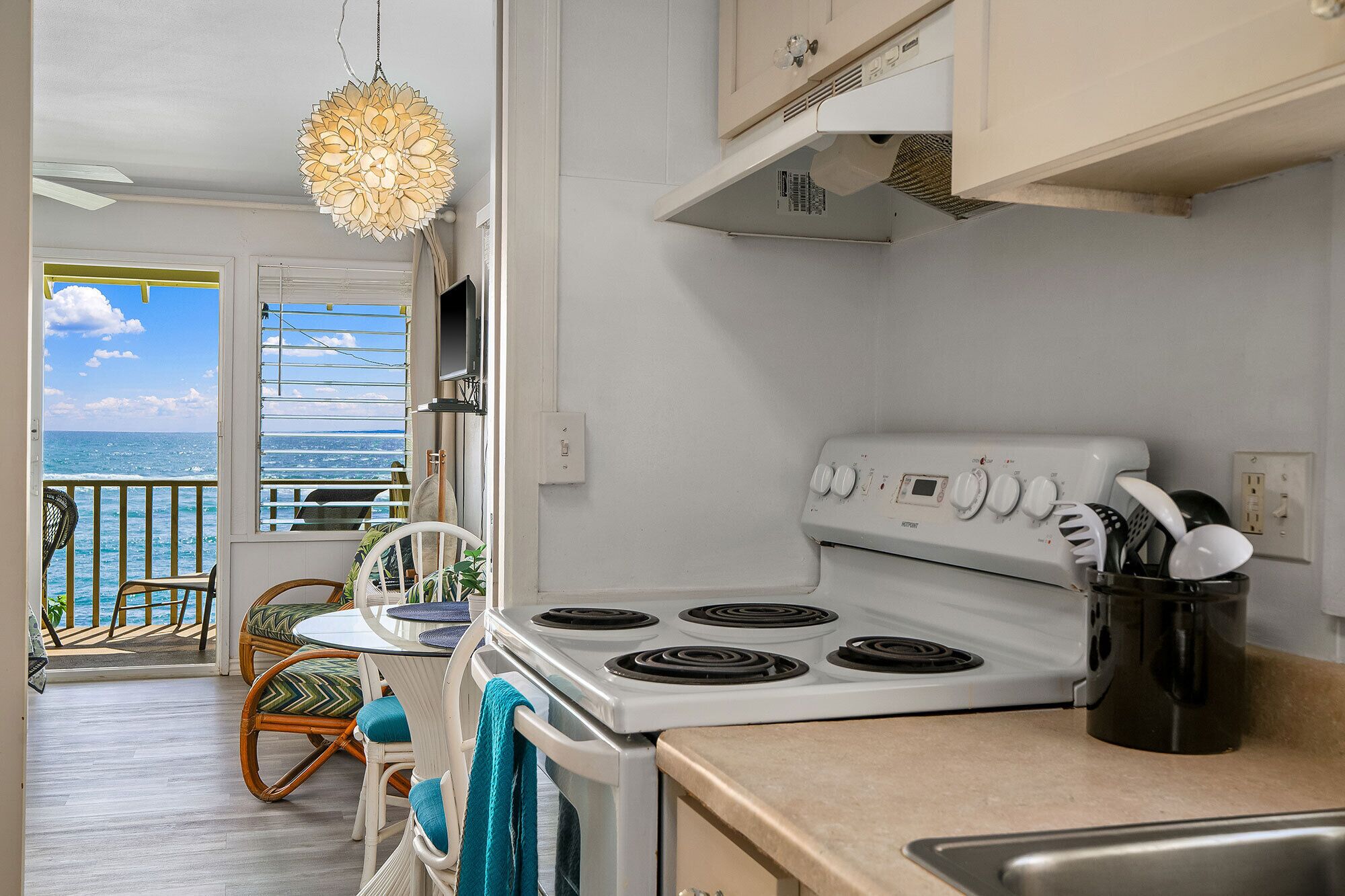 A kitchen with a white stove, a dining table with chairs, a hanging light fixture, and a view of the ocean through a window.