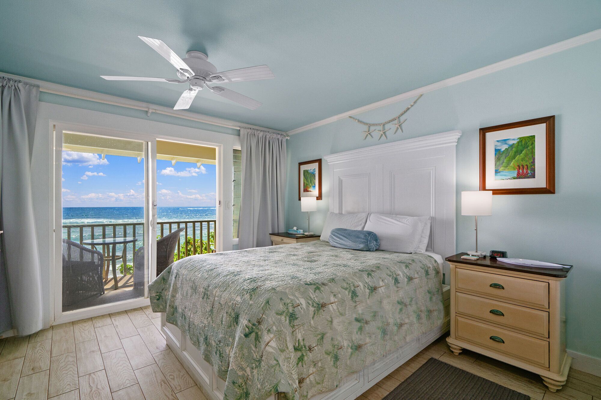 A bedroom with a bed, a nightstand, a ceiling fan, a window with a view of the ocean, and a door leading to a balcony.
