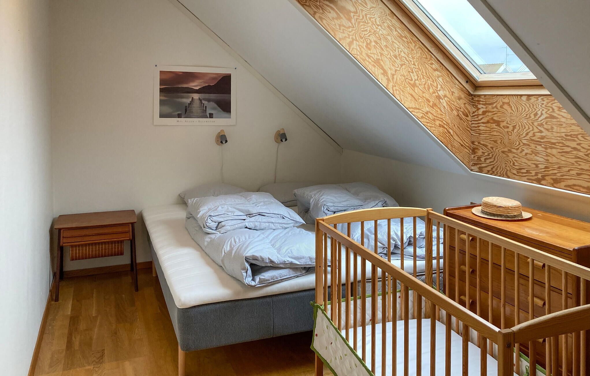 Een kleine kamer met een schuin dak, een hemelbed, een houten kinderbedje, een tafeltje en een lijst met een afbeelding aan de muur.