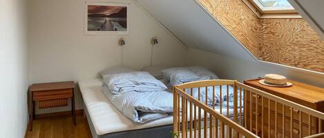 Een kleine kamer met een schuin dak, een hemelbed, een houten kinderbedje, een tafeltje en een lijst met een afbeelding aan de muur.