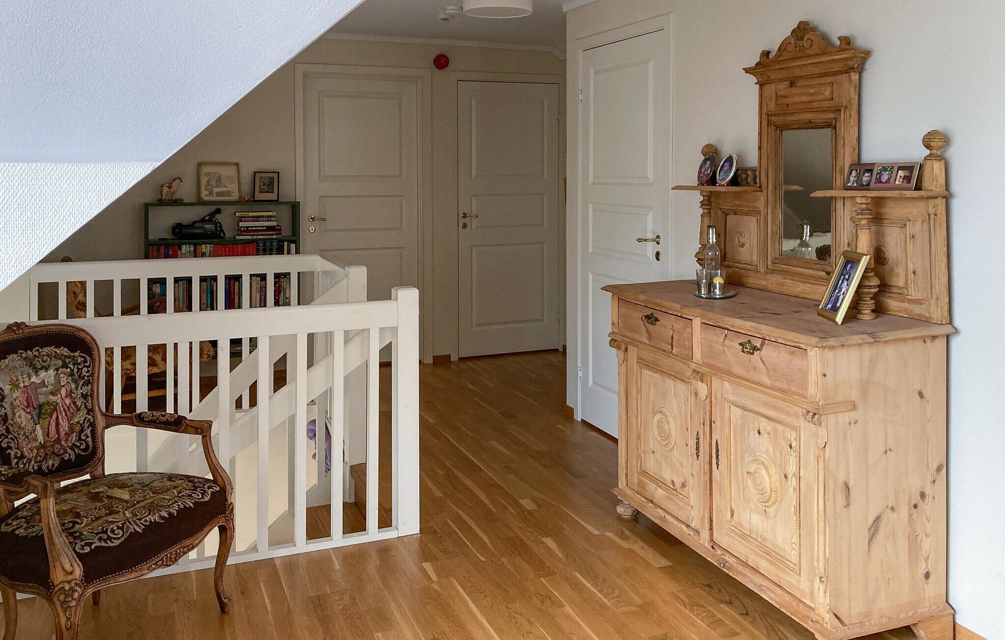 Ett rum med trägolv, en vit dörr, en byrå i trä med en spegel och en stol med ett blommigt sittkudd.