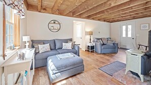 A spacious living room with a high wooden ceiling, a grey sofa set, a fireplace, and a dining table with chairs.