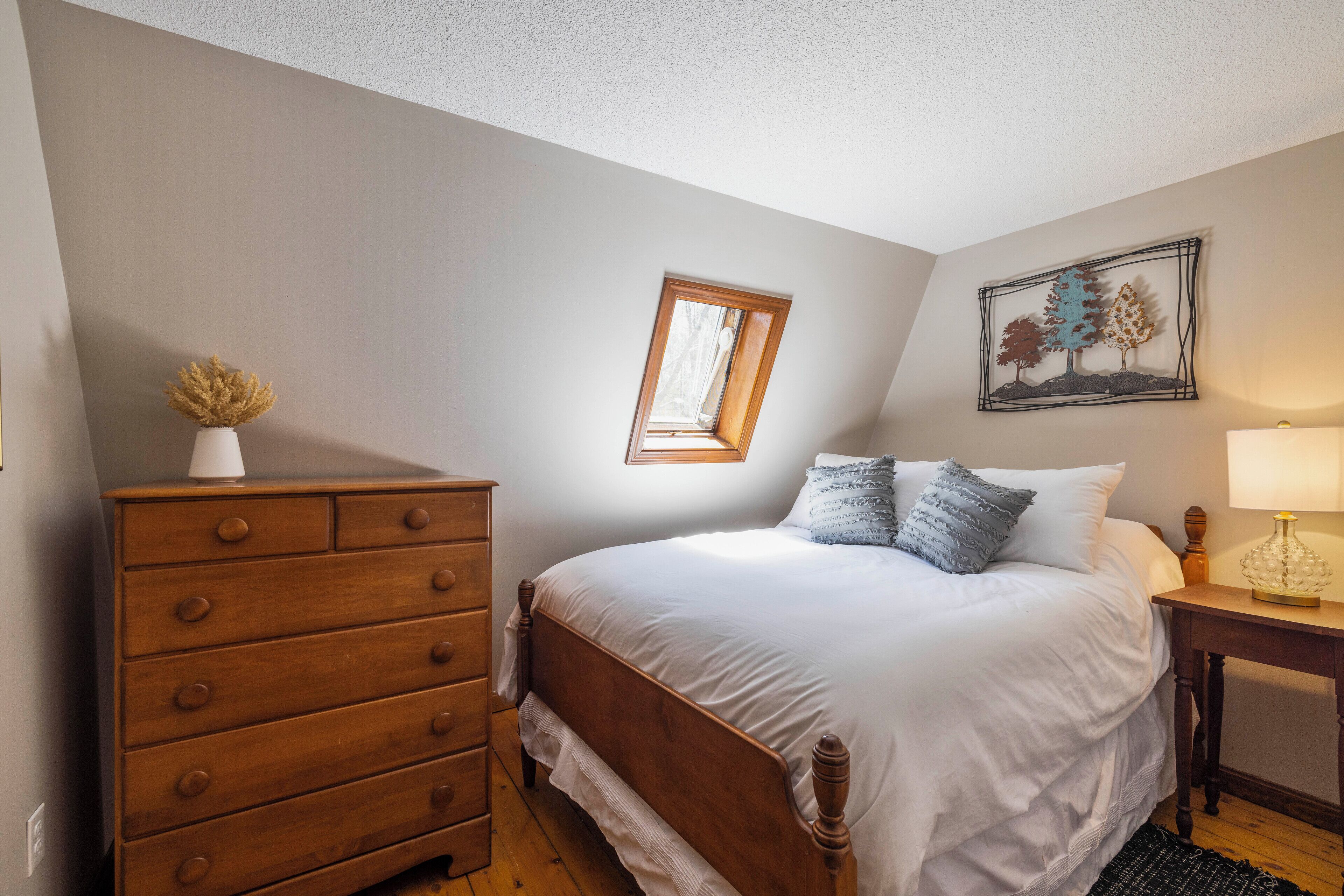 A bedroom with a wooden dresser, a bed with white bedding, a small window, and a wall-mounted picture.