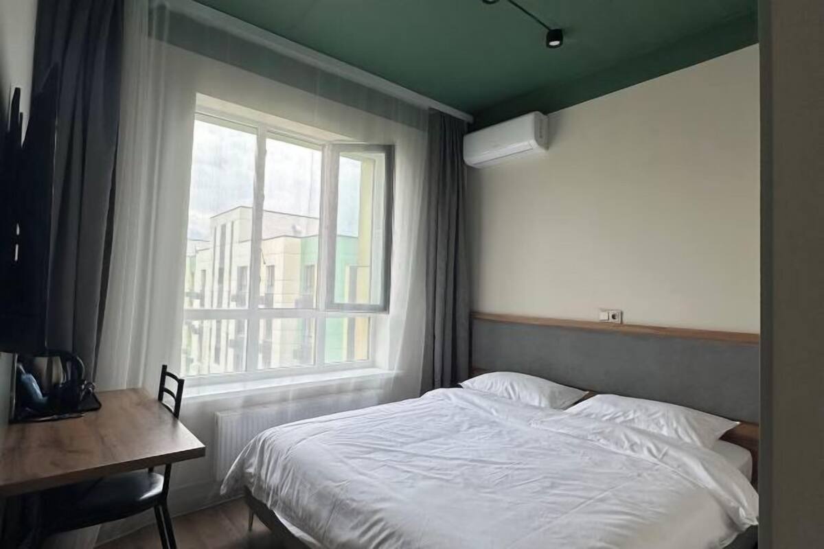 A hotel room with a bed, a desk with a chair, a window with curtains, and an air conditioning unit on the wall.