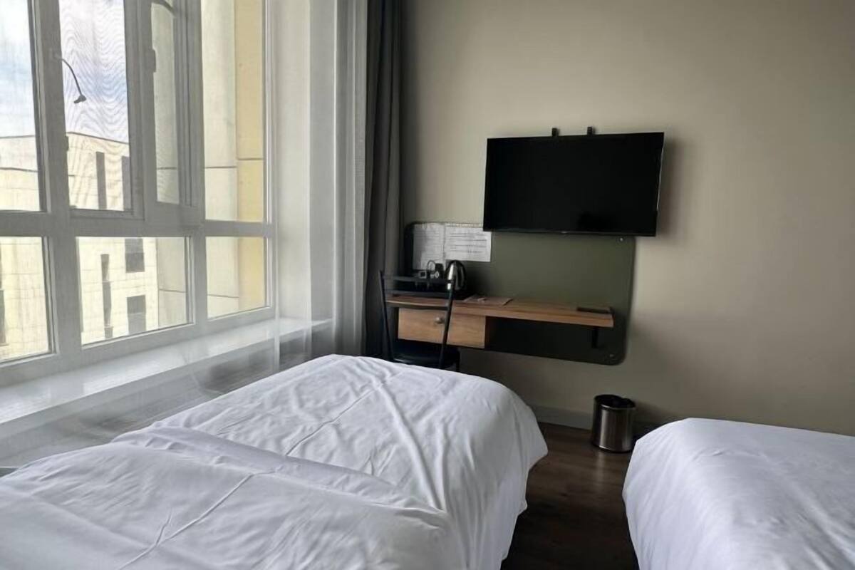 A hotel room with two beds, a desk with a flat-screen TV, a window with a view of buildings, and a trash can.