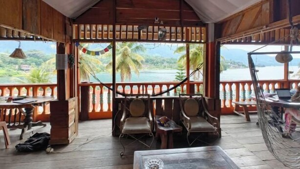A wooden veranda with a view of palm trees and a body of water. There are wooden chairs and a table. - Sibayak Guest house (Samosir Regency)