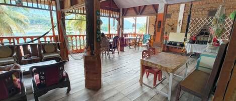 A wooden-floored room with a table and chairs, a hammock, and a view of the sea.