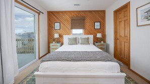 Une chambre avec un grand lit, un ventilateur de plafond, des murs recouverts de panneaux de bois, une fenêtre avec vue et une porte.