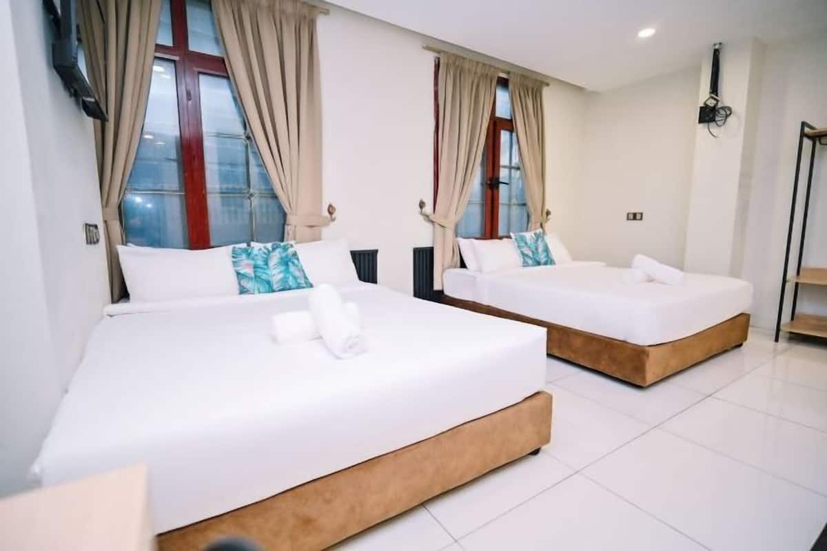 Two single beds with white bedding and wooden bases in a room with large windows and beige curtains.