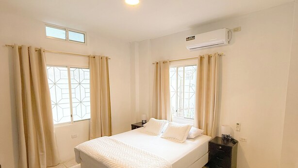 Quarto limpo e bem iluminado com cama, cortinas, mesa de cabeceira, aparelho de ar condicionado e um vaso de planta.