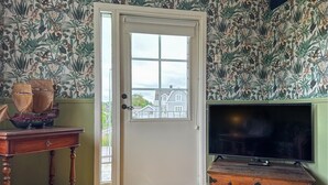 A room with a door, a wooden table with a ship model, a television on a wooden cabinet, and a wall with a tropical wallpaper pattern. - 2 bedroom beautiful home in Påskallavik (Påskallavik)