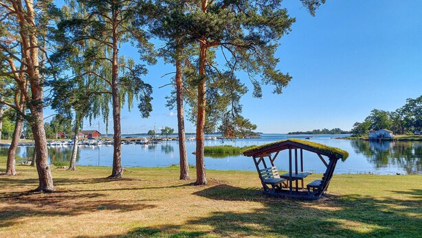 A lakeside gazebo with a checkered seat, surrounded by tall trees and a clear view of boats on the water. - 2 bedroom beautiful home in Påskallavik (Påskallavik)
