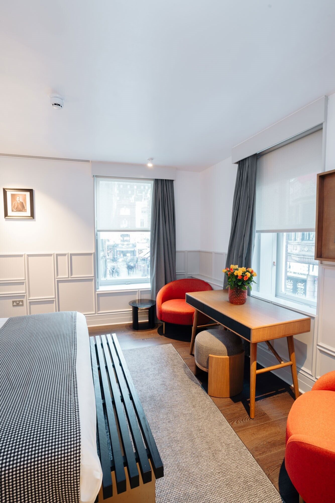 A modern hotel room with a bed, a desk, two chairs, a small table with flowers, and large windows.