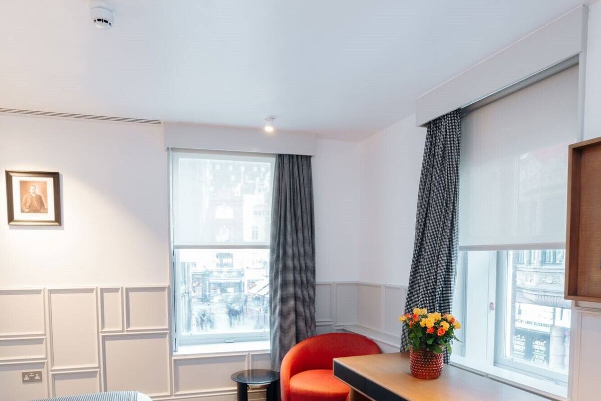 A modern hotel room with a bed, a desk, and two chairs. There is a window with a view of buildings outside.