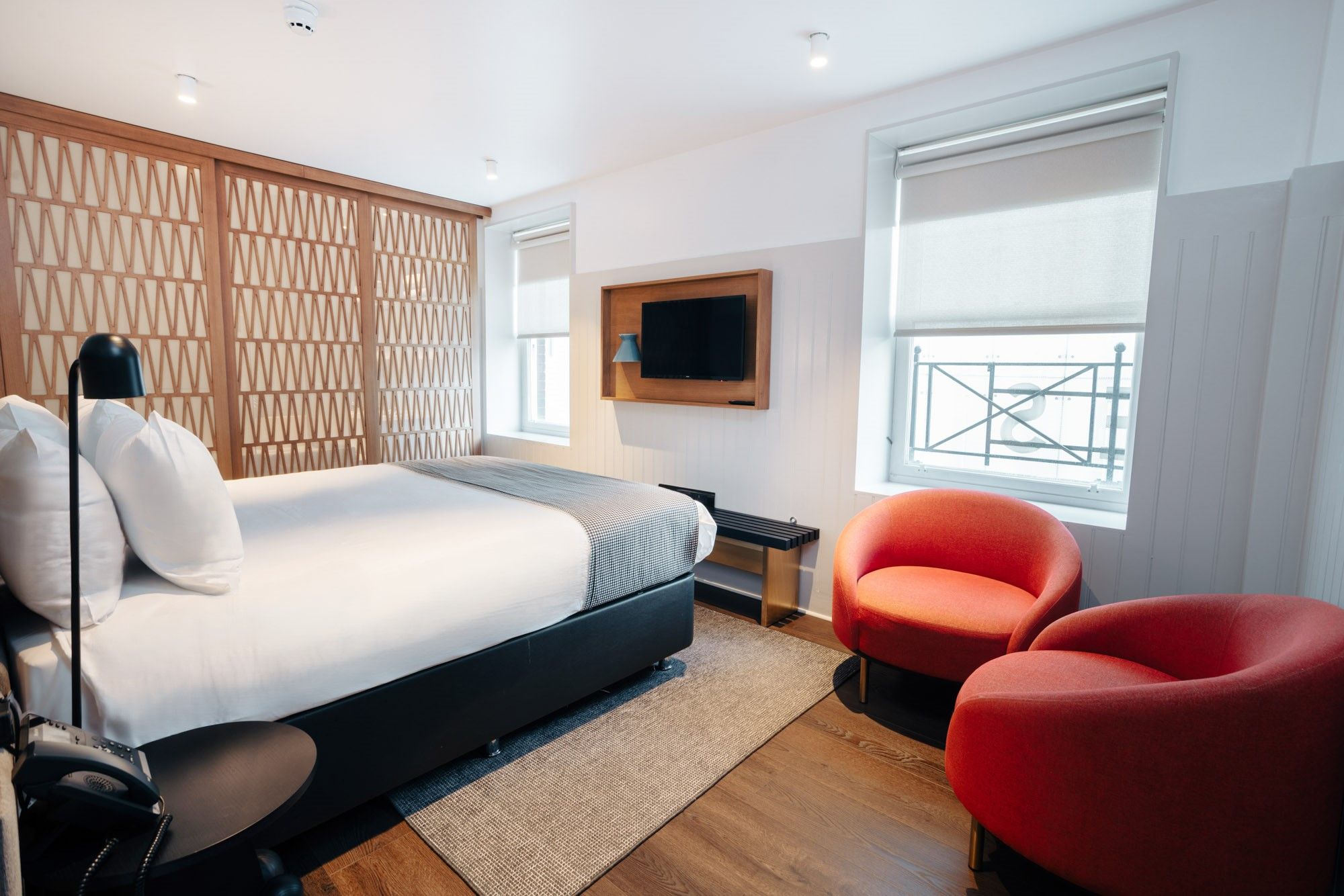 A modern hotel room with a bed, a TV mounted on the wall, and two red chairs.