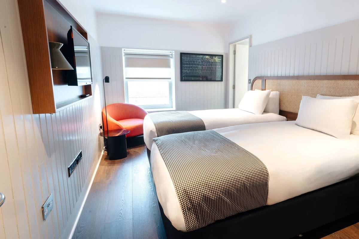 A modern hotel room with two beds, a TV mounted on the wall, a window with a view, and a door leading to the bathroom.