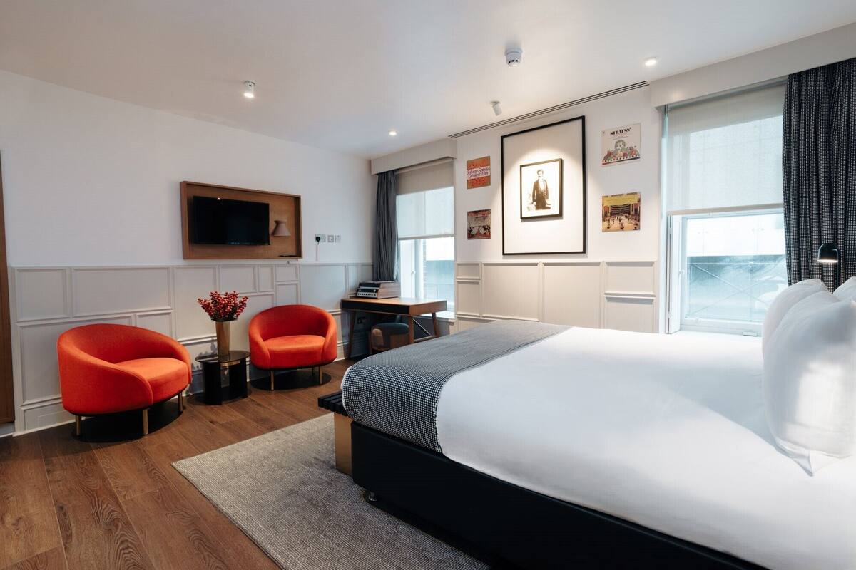 A modern hotel room with a large bed, two red armchairs, a desk with a flat-screen TV, and artwork on the walls.