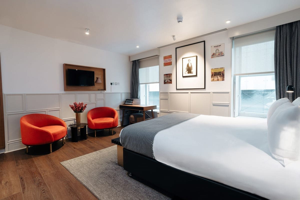 A modern hotel room with a large bed, two red armchairs, a desk with a flat-screen TV, and artwork on the walls.