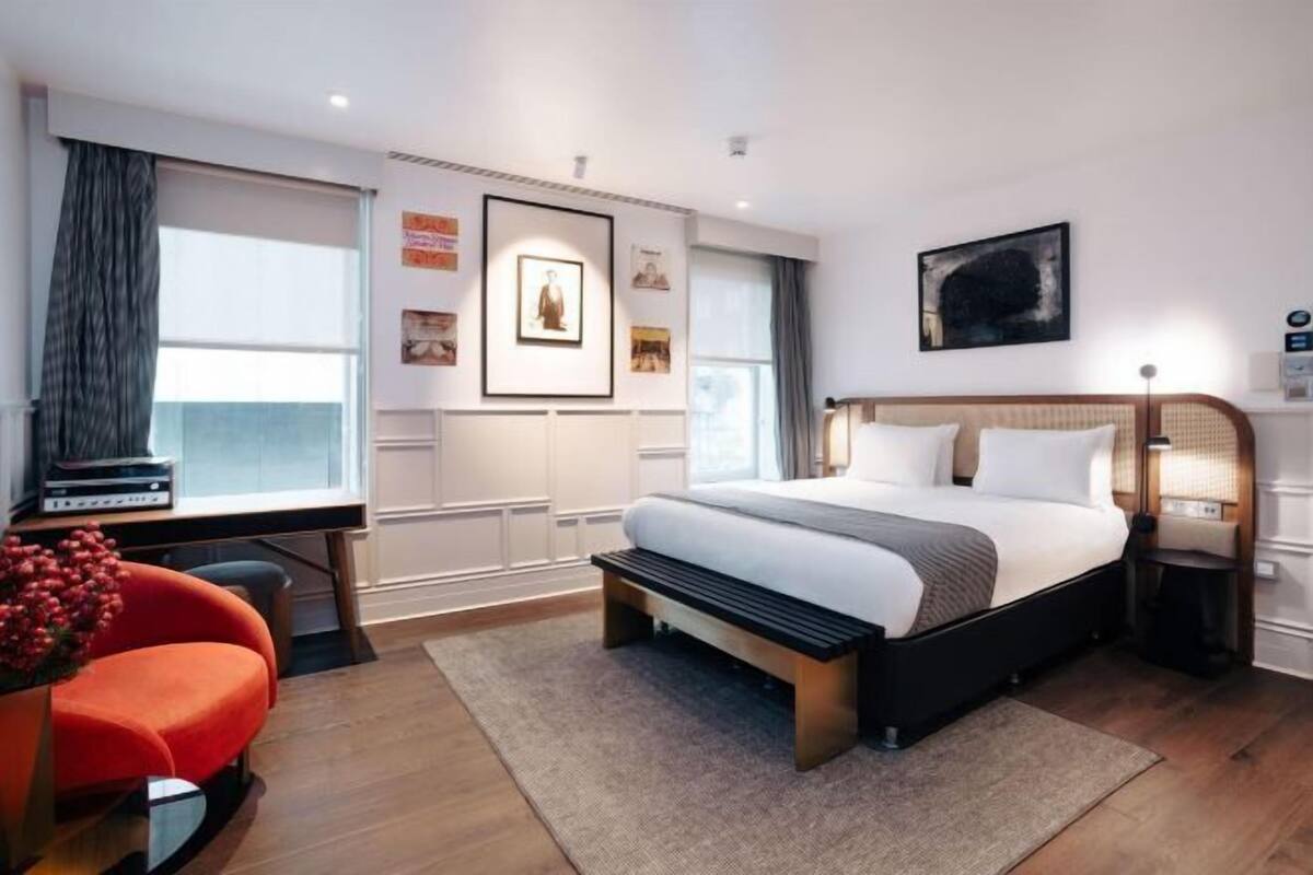 A modern hotel room with a large bed, a desk, and a chair. There are framed pictures on the wall and a window with curtains.
