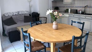 En kompakt opholdsstue med spisebord og stole, en sofa med hynder, et tekøkken med skabe og hårde hvidevarer, samt en vase med blomster på bordet.