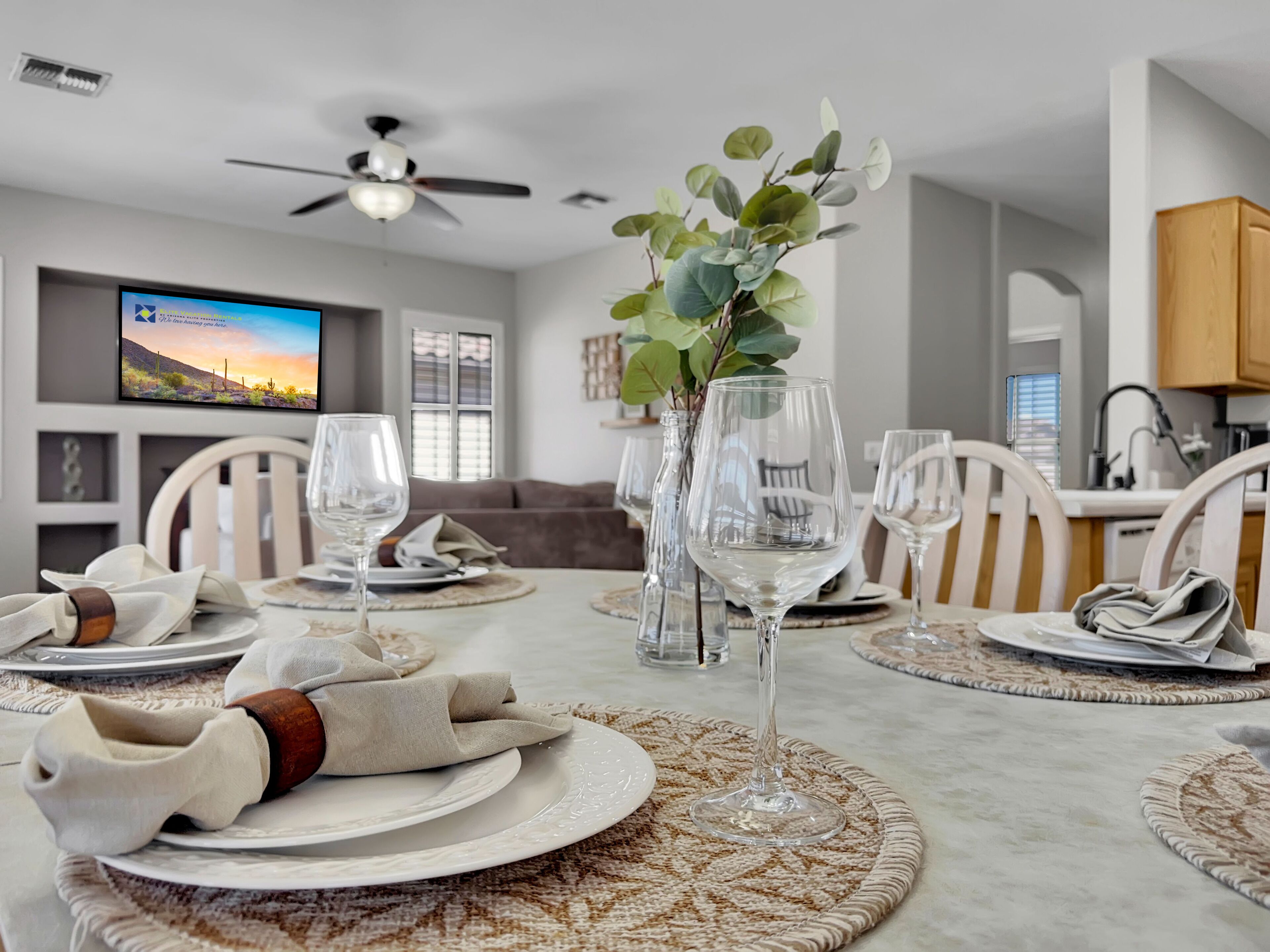 A dining area with a table set for six, including wine glasses, plates, and cutlery. There is a television mounted on the wall displaying a scenic image.
