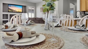 Uma sala de jantar com uma mesa posta para seis pessoas, incluindo copos de vinho, pratos e talheres. Há uma televisão na parede exibindo uma imagem panorâmica.