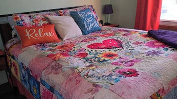 Een bed met een quilt met bloemenprint, een rood kussen met de tekst 'Relax' en een blauw kussen met de tekst 'Home Sweet Home'.