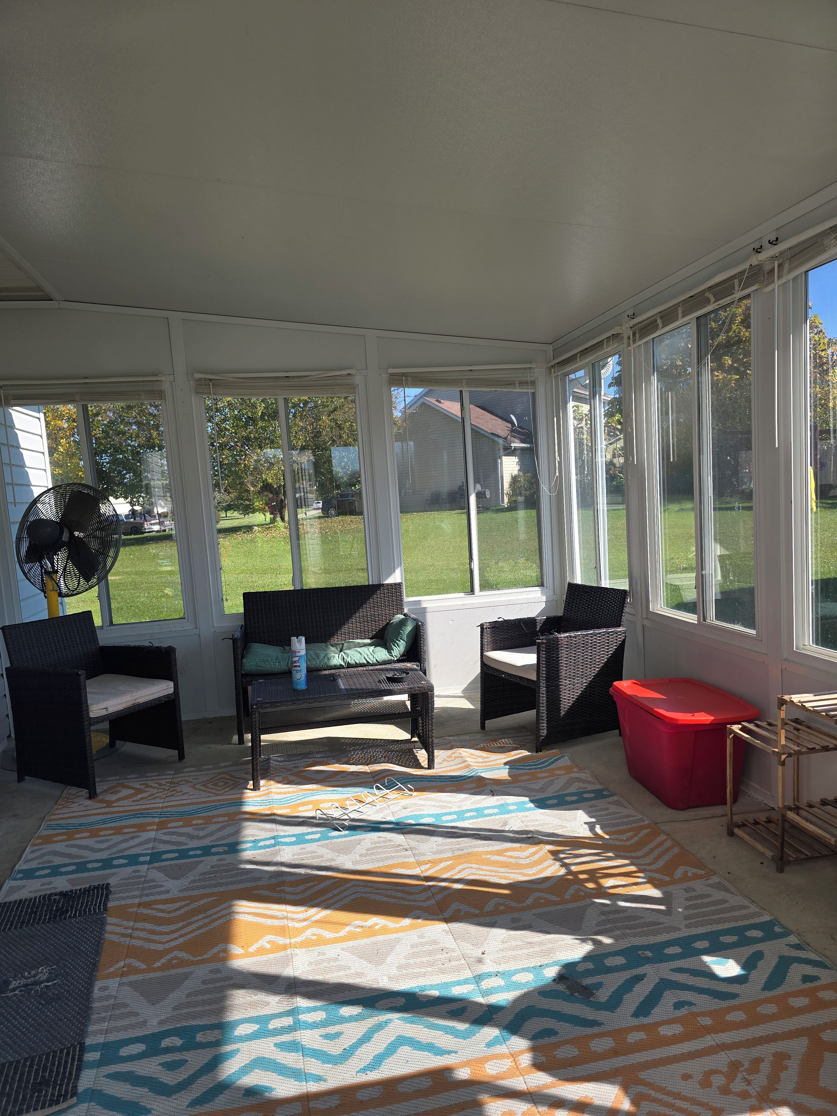 Een zonnig terras met rieten meubilair, een rode opbergkist en een vloerkleed met dessin.