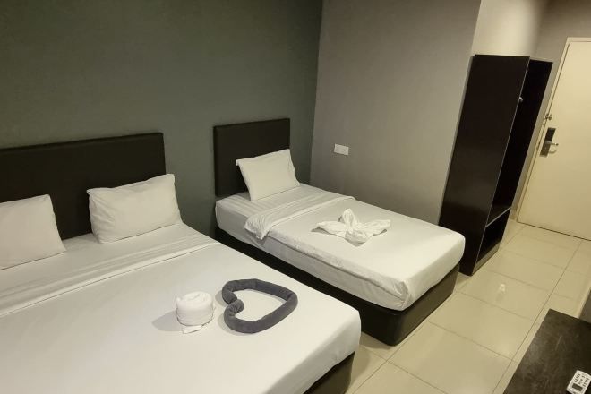 A hotel room with two beds, each with white bedding and pillows, a dark headboard, and a small table with a white cup and a heart-shaped pillow.