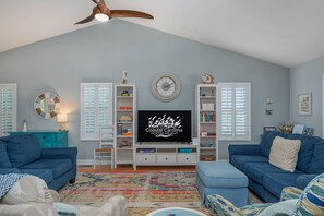 A living room with a television displaying 'Coastal Carolina Vacations and Sales', two blue sofas, a colorful rug, and a ceiling fan.