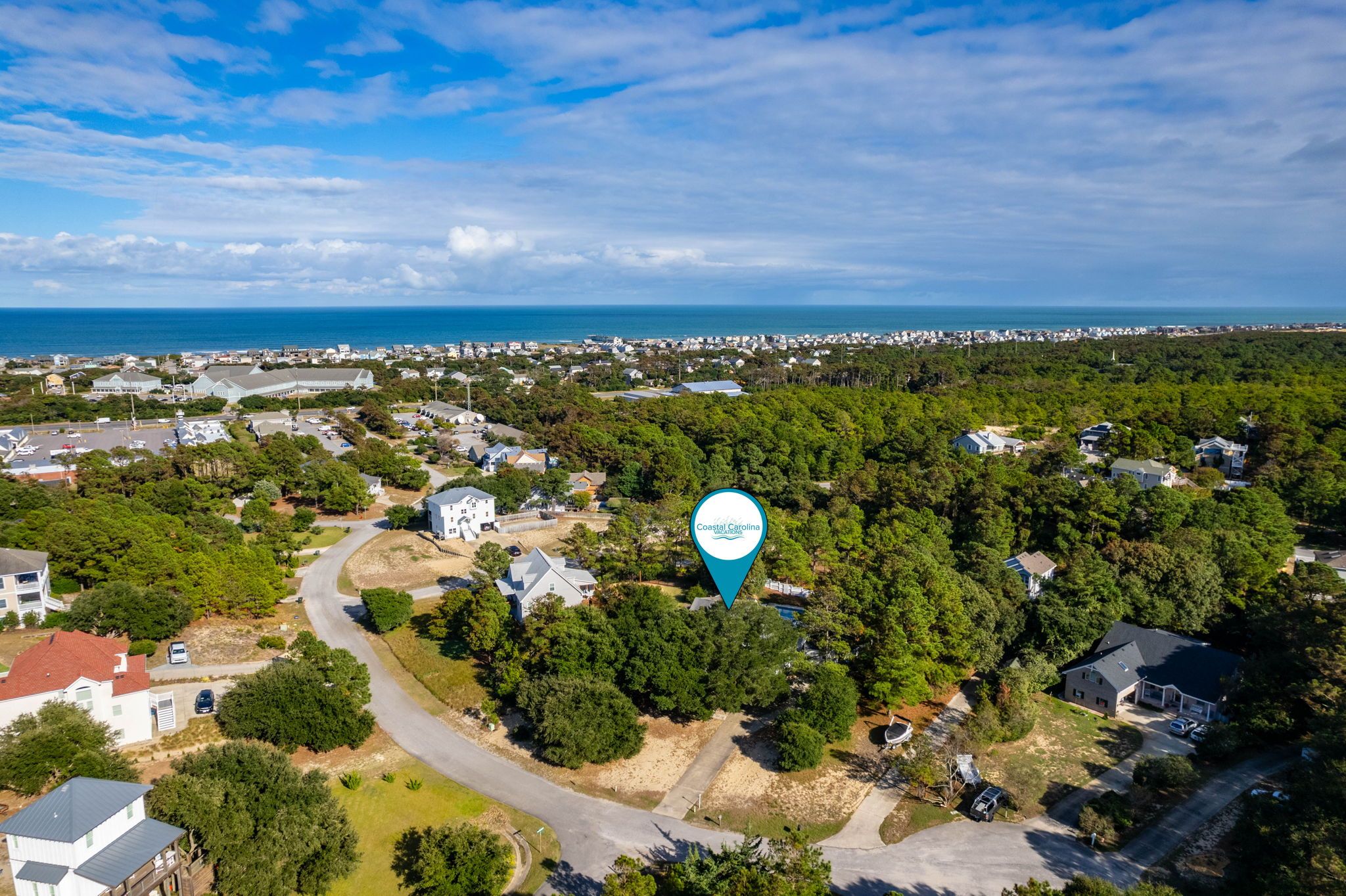 An aerial view of a coastal residential area with houses, a road, and a body of water in the background.