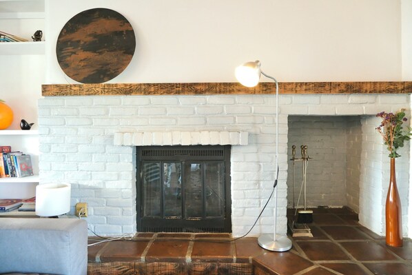 Un salón con chimenea de ladrillo blanco, un adorno circular en la pared, una lámpara de pie y una estantería con libros y objetos decorativos.