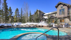 A swimming pool with a hot tub, lounge chairs, and a building with a ski lodge in the background.