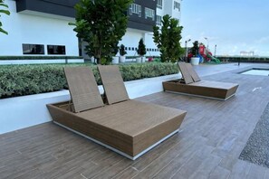 Dos sillones con cojines, suelo de baldosas y un edificio con ventanas y vegetación al fondo.