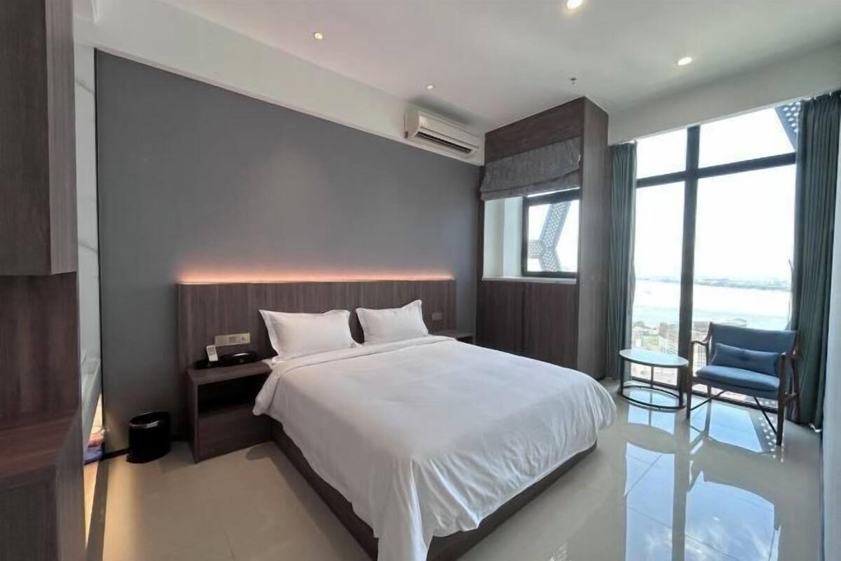 A modern hotel room with a large bed, a small table, a chair, and a view of the outdoors.