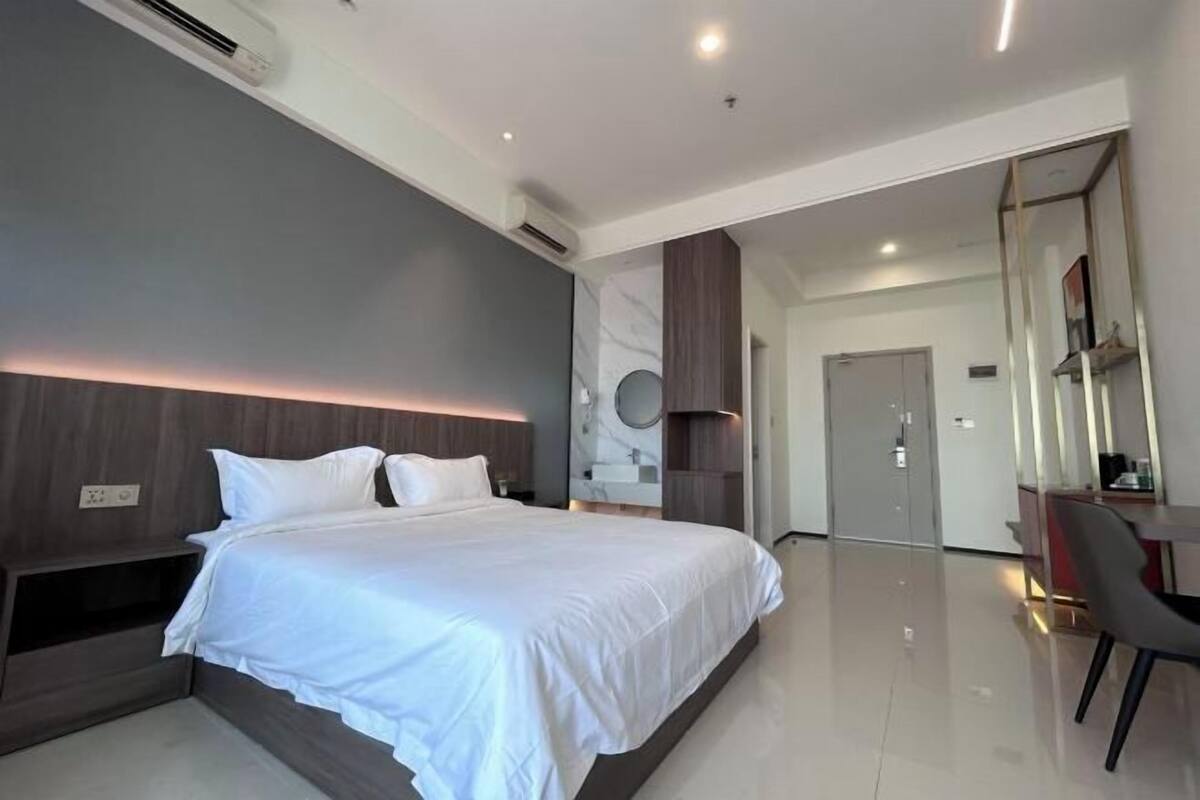 A modern hotel room with a large bed, a desk with a chair, a mirror, and a wardrobe.