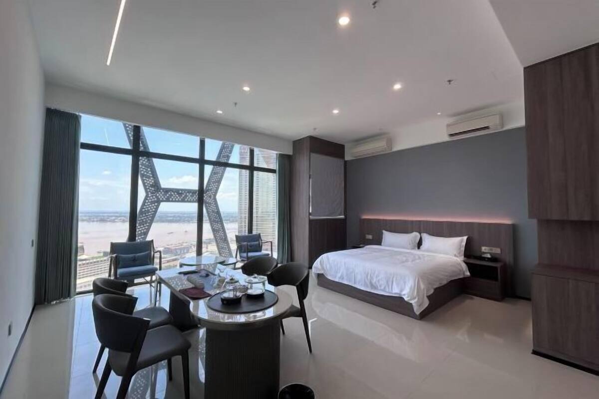 A modern hotel room with a large bed, a dining table with chairs, and a view of the cityscape.